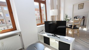 A modern living room with a flat-screen TV on a white console, a round side table, a dining area with a wooden table and chairs, and a view of buildings through a window. - FullHouse | Chemnitz | M45-2 | Studio (Chemnitz)