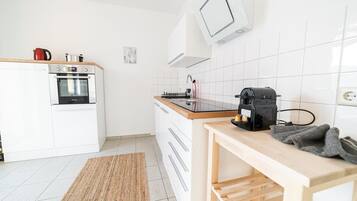 A modern kitchen with white cabinets, a built-in oven, a coffee maker, and a wooden table.