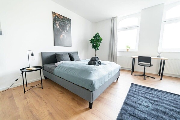 Un dormitorio moderno con una cama gris, una mesita de noche pequeña, un escritorio con una silla, una planta en maceta y una ventana con cortinas.