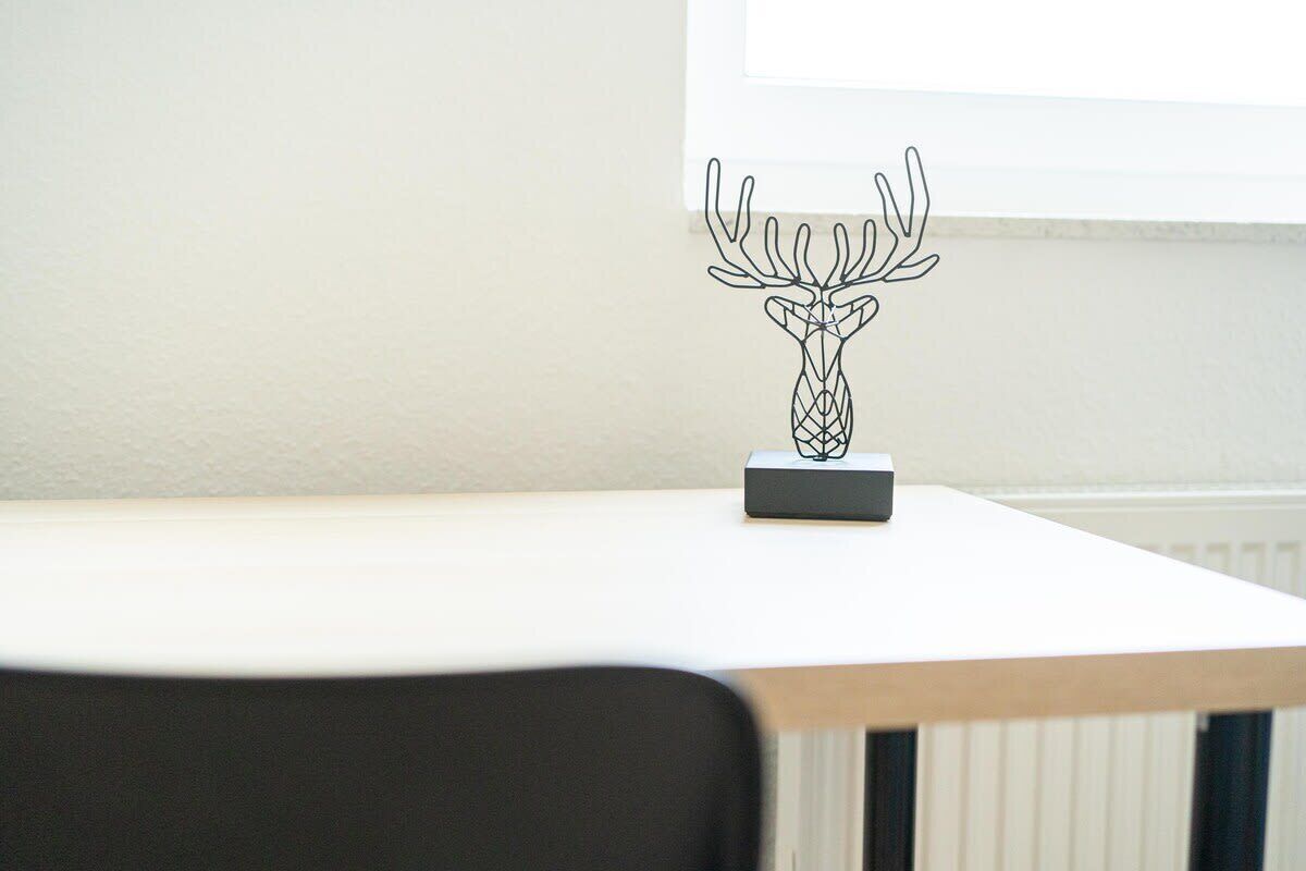 Een witte tafel met een zwarte stoel ervoor, een zwart voorwerp op de tafel, en een raam op de achtergrond.