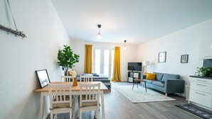 A modern living room with a dining area, a grey sofa, a TV, and a plant. - FULL HOUSE Premium Apartment Dresden (Dresden)
