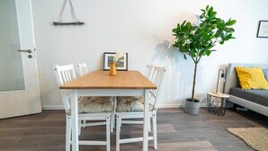 A dining area with a wooden table and white chairs, a potted plant, and a gray sofa with a yellow pillow. - FULL HOUSE Premium Apartment Dresden (Dresden)