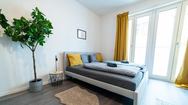 A modern bedroom with a bed, a potted plant, a small side table, and a window with curtains. - FULL HOUSE Premium Apartment Dresden (Dresden)