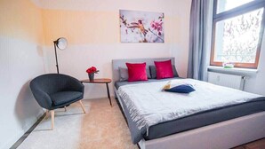 Un dormitorio con cama, una silla, una mesita con un florero y una ventana con vistas.
