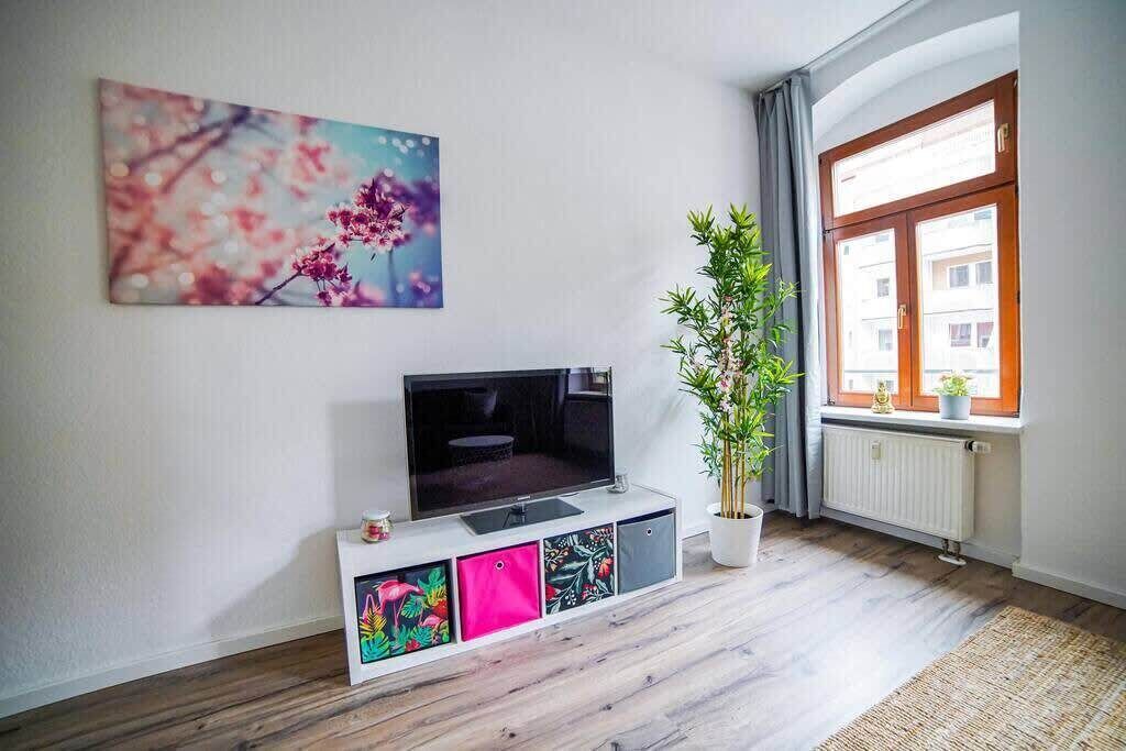 Een woonkamer met een flatscreen-tv op een witte console, een potplant, een raam met uitzicht op gebouwen en een bloemen schilderij aan de muur.