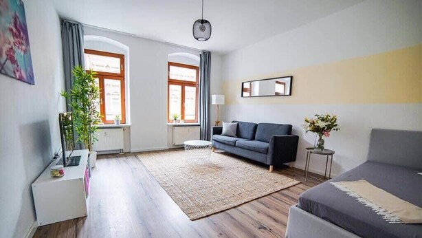 Una sala de estar moderna con un sofá gris, una pequeña mesita auxiliar, un ventanal con cortinas, una lámpara de pie y una planta en maceta.