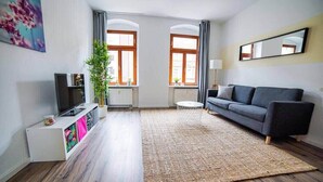 Una sala moderna con un sofá gris, un televisor de pantalla plana sobre un mueble blanco, un tapete tejido grande y dos ventanas con cortinas.