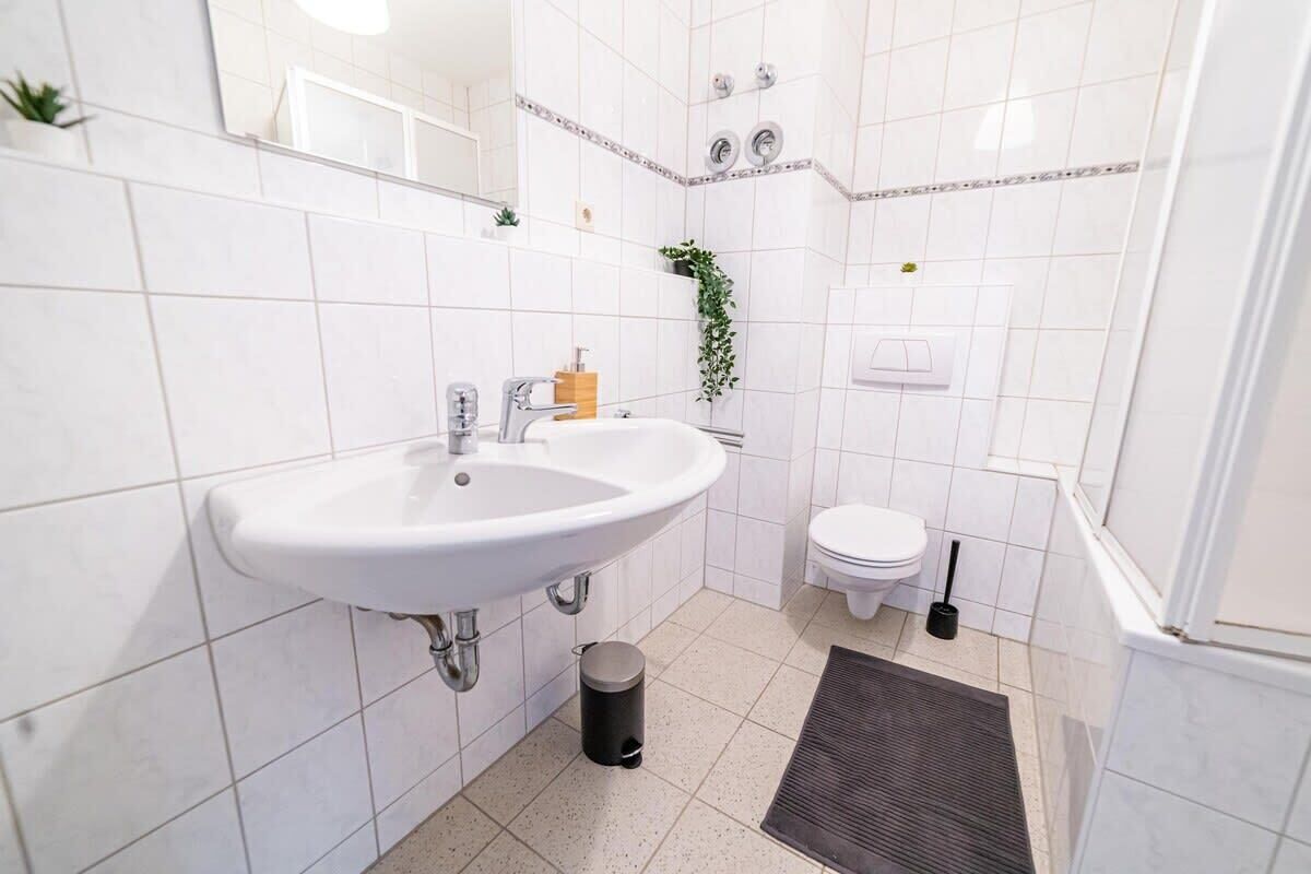 Un baño con lavamanos, inodoro y ducha blancos. Hay un cubo de basura y una pequeña planta en la pared.