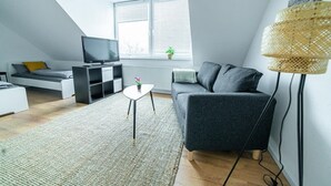 A modern living room with a sofa, a TV, a small table with a plant, and a window with blinds. - FullHouse | Magdeburg | L10-5 | RoofTop (Magdeburg)