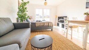En moderne stue med en grå sofa, et rundt sort sofabord, et spisebord med stole, et fjernsyn og et stort vindue med udsigt over det fri.