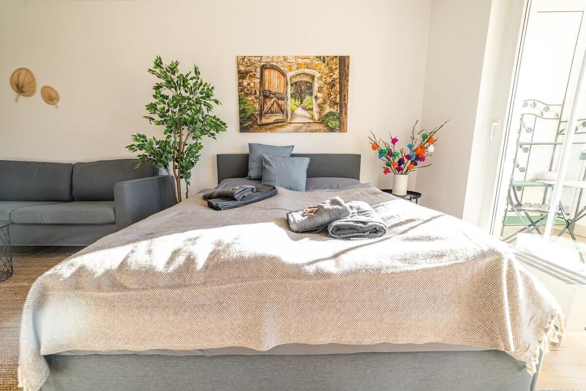 Ein Schlafzimmer mit einem Bett, einem grauen Sofa, einer Topfpflanze, einem Gemälde und einer Vase mit Blumen.