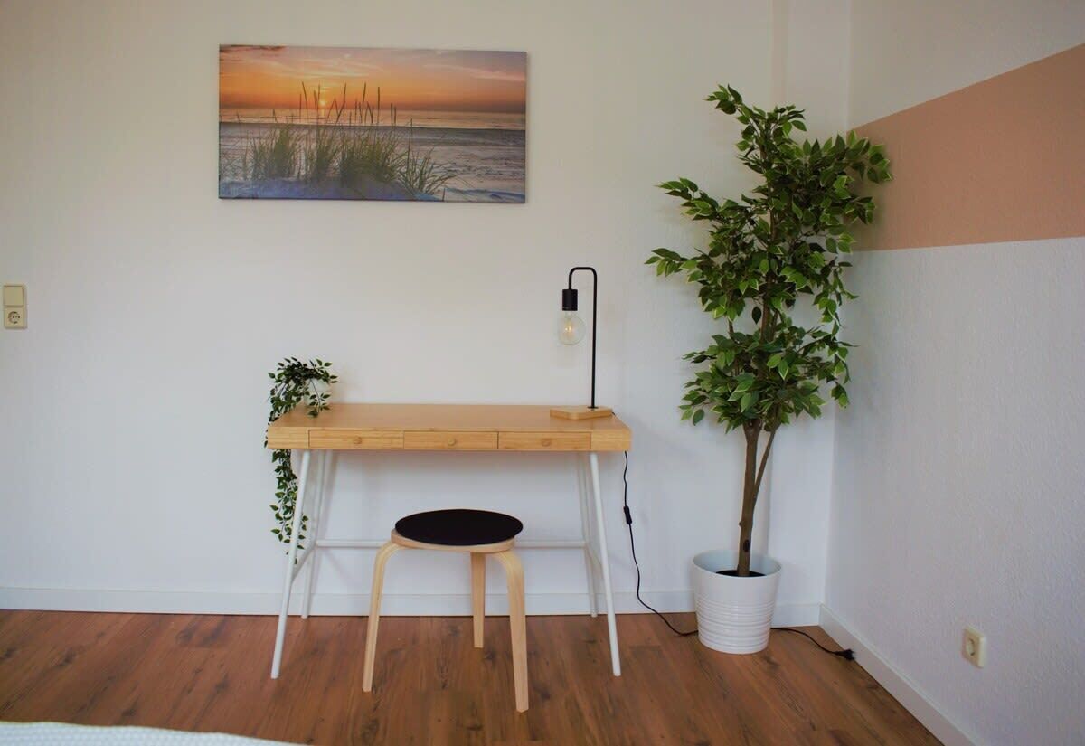 A room with a wooden desk, a black chair, a potted plant, and a wall-mounted lamp.
