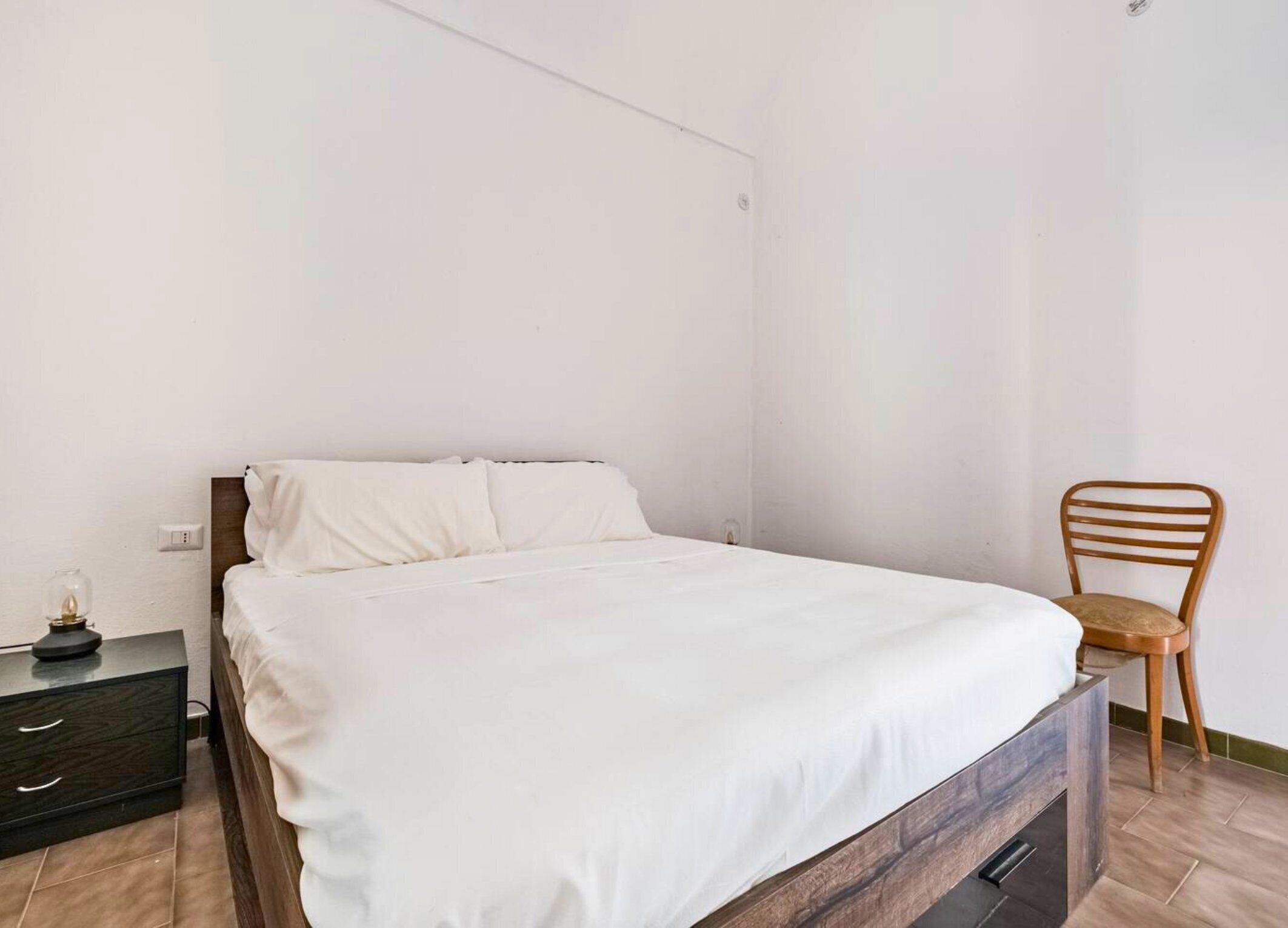 A neatly made bed with white linens in a simple room with a wooden chair and a small bedside table.
