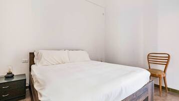 A neatly made bed with white linens in a simple room with a wooden chair and a small bedside table.
