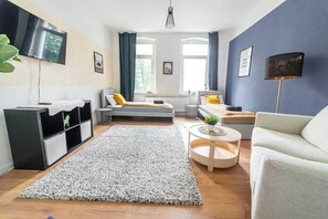 Een moderne woonkamer met een bank, een ronde salontafel, een aan de muur bevestigde televisie en een groot raam met gordijnen.