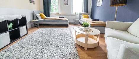 Una sala de estar moderna con sofá, una mesita redonda, una cama con cabecera y una ventana con cortinas.