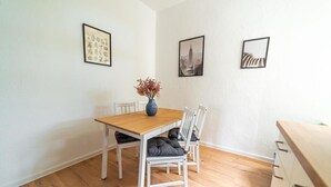 Un comedor con una mesa de madera y sillas blancas, un florero con flores secas y dos cuadros en la pared.