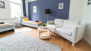 Una sala de estar moderna con un sofá, una mesita redonda pequeña, una lámpara de pie y una cama con un almohadón amarillo.