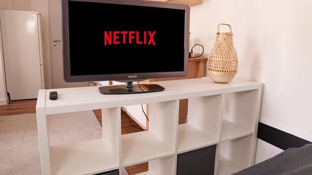 A television on a white stand with the word "NETFLIX" on the screen, a remote control, and a woven lantern on the stand. - FullHouse | Halle | T28 Panda Apt. (Halle (Saale))
