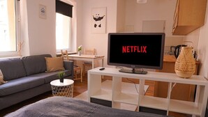 A modern living room with a grey sofa, a small round table with a plant, a white TV stand displaying a Netflix logo, and a wooden dining table with chairs. - FullHouse | Halle | T28 Panda Apt. (Halle (Saale))