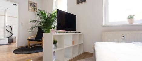 A modern bedroom with a bed, a TV on a white stand, a potted plant, and a window with a view.