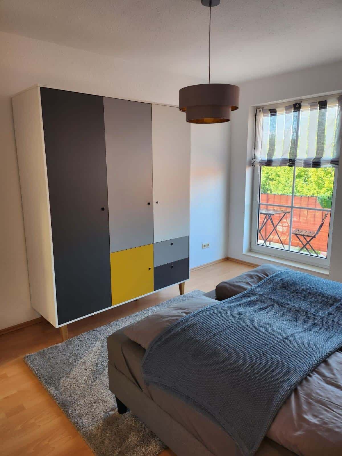 A bedroom with a bed, a grey and yellow wardrobe, a hanging lamp, and a window with a view of a patio.