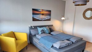Una camera da letto con un letto, una poltrona gialla, un quadro raffigurante un tramonto, una lampada e uno specchio a parete.
