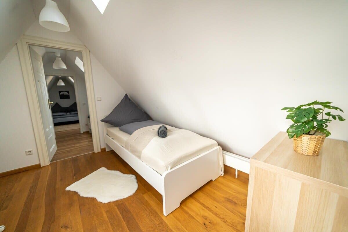 A small, single-bed room with a sloped ceiling, a wooden floor, a white bed with a gray pillow, a white rug, a small table with a plant, and a white lamp.