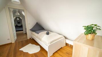 A small, single-bed room with a sloped ceiling, a wooden floor, a white bed with a gray pillow, a white rug, a small table with a plant, and a white lamp.