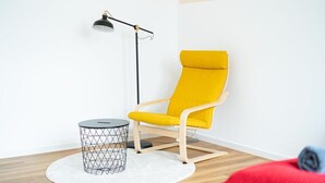 A room with a yellow chair, a small round table with a wireframe base, and a floor lamp. - FullHouse | Magdeburg | L10-2 | FamilienApartment (Magdeburg)