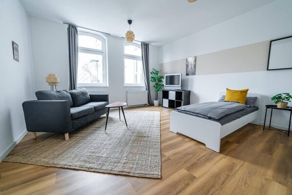 Ein modernes Schlafzimmer mit einem grauen Sofa, einem weißen Bett, einem kleinen Tisch und einem Fernseher.