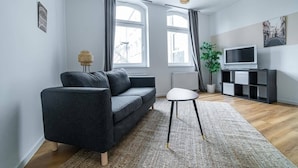 A modern living room with a dark grey sofa, a white side table, and a large window with curtains. - FullHouse | Magdeburg | L10-2 | FamilienApartment (Magdeburg)