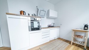 A modern kitchen with white cabinetry, a built-in oven, a coffee machine, and a small wooden side table. - FullHouse | Magdeburg | L10-2 | FamilienApartment (Magdeburg)