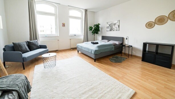 A modern bedroom with a bed, a grey sofa, a small side table, and a large window.