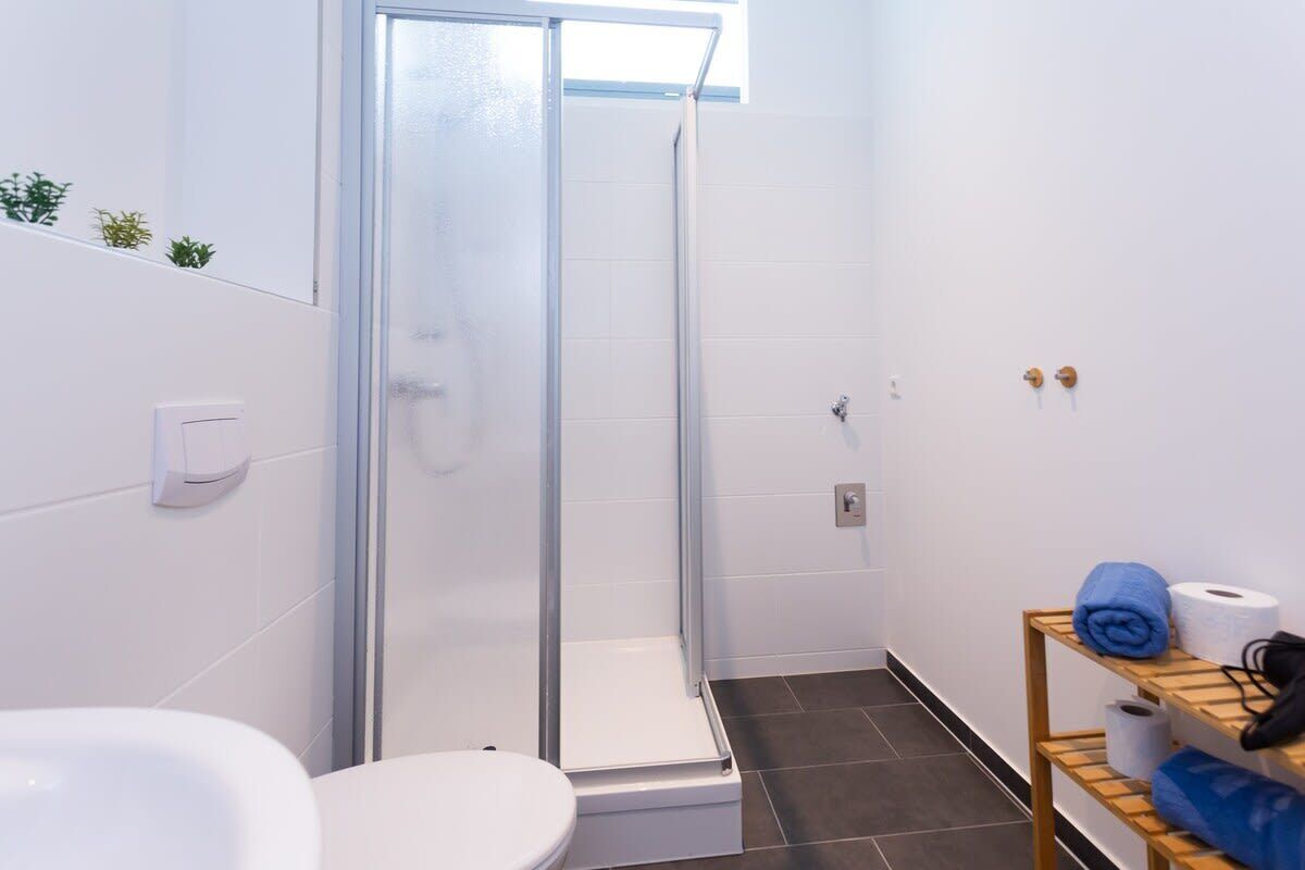A bathroom with a glass shower enclosure, a white toilet, a sink, and a shelf with towels and toiletries.