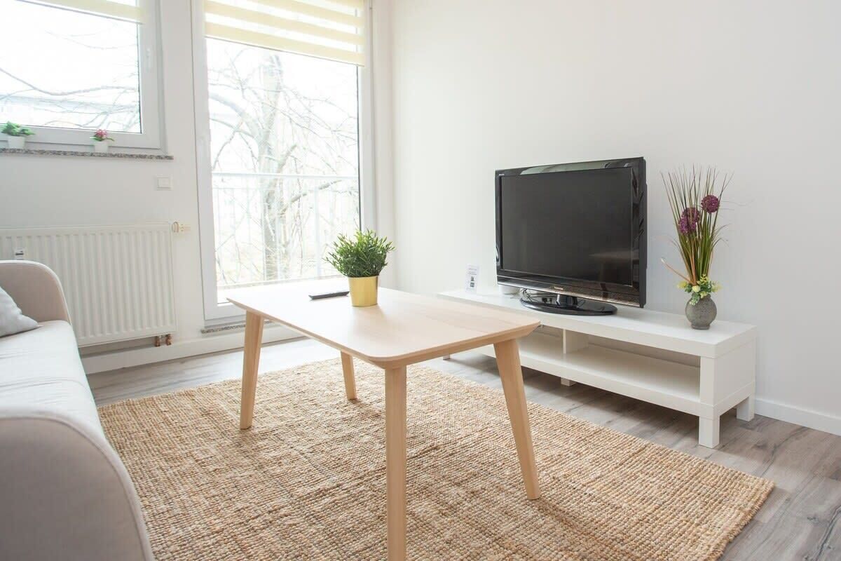Un salon moderne avec un canapé, un téléviseur posé sur un meuble blanc, une petite table et une fenêtre donnant sur des arbres.