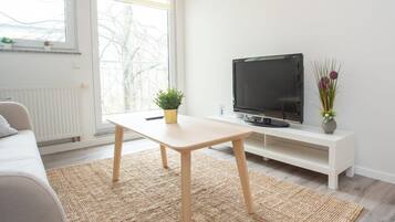 Un salon moderne avec un canapé, un téléviseur posé sur un meuble blanc, une petite table et une fenêtre donnant sur des arbres.