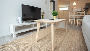 Un salon moderne doté d’un téléviseur à écran plat, d’une console blanche, d’une petite plante en pot et d’un coin repas avec des chaises et une table en bois.