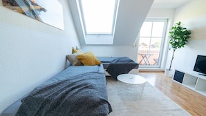 Ein modernes Schlafzimmer im Loft-Stil mit Dachfenster, einer Schlafsofa, einem runden Couchtisch und einem Fernseher auf einem weißen Lowboard.