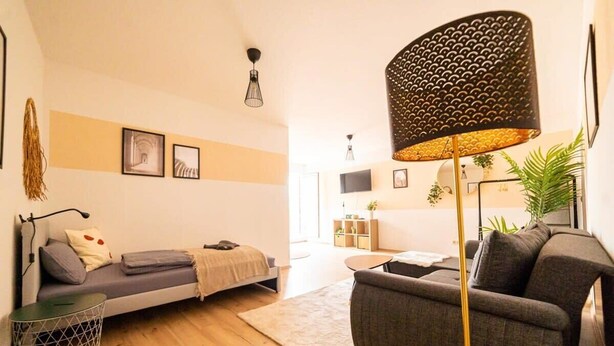 Ein modernes Wohnzimmer mit Sofa, Sessel und Stehlampe. Ein kleiner Beistelltisch und eine Zierpflanze sind ebenfalls vorhanden.
