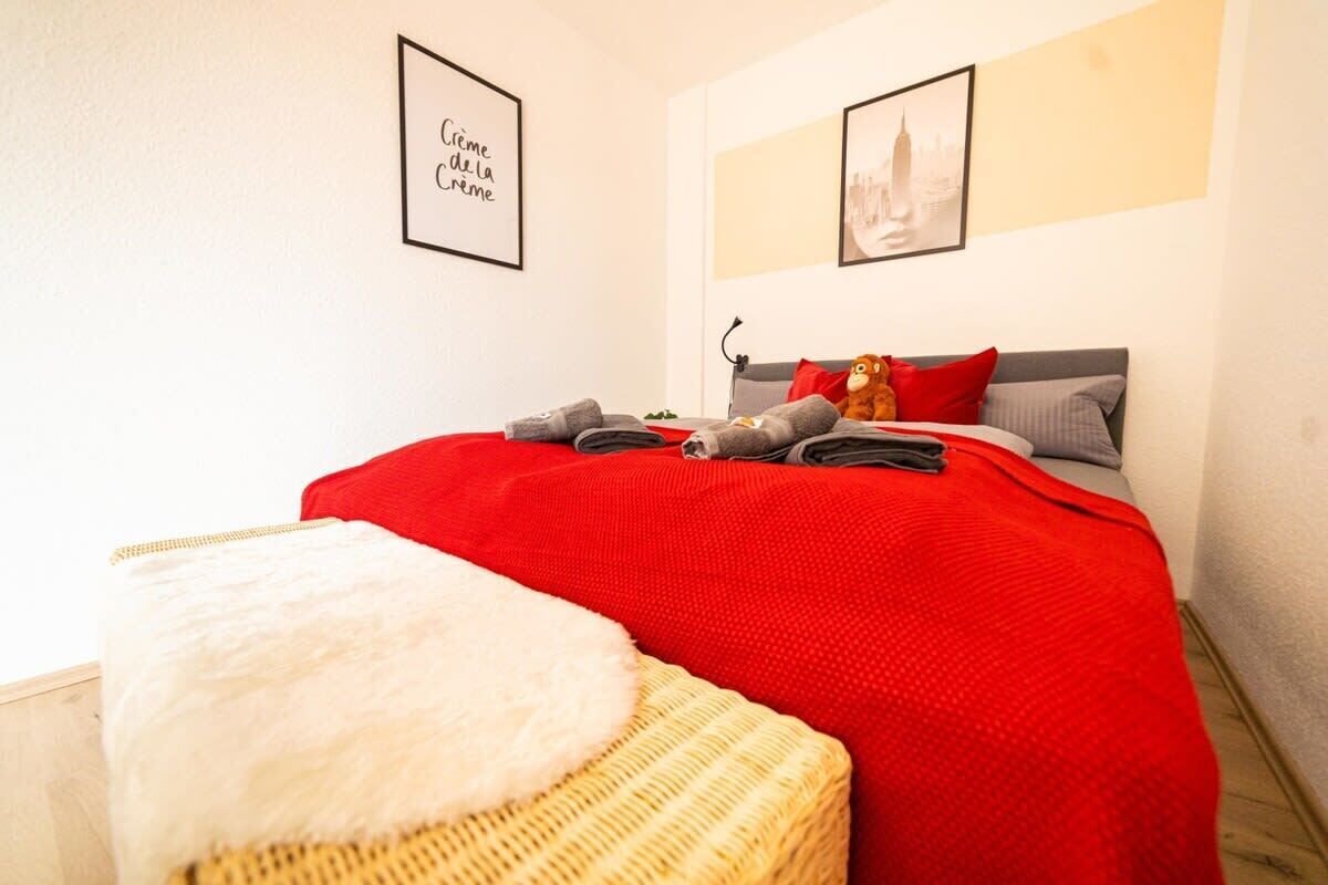 Un lit bien fait, recouvert d’une couverture rouge et agrémenté d’oreillers, dont un oreiller blanc et moelleux, ainsi qu’un petit tabouret en osier. Deux tableaux encadrés ornent le mur, l’un avec du texte et l’autre représentant un bâtiment.