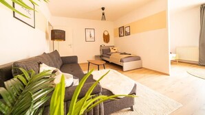 Ein modernes Wohnzimmer mit einem grauen Sofa, einem kleinen grauen Zweisitzer und einem Couchtisch. An der Wand befinden sich gerahmte Bilder und auf einem Beistelltisch steht eine Lampe.