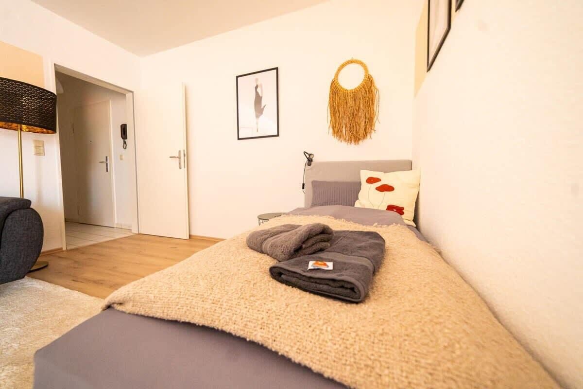A bedroom with a bed, a bedside table, a wall-mounted lamp, a framed picture, and a decorative wall hanging.