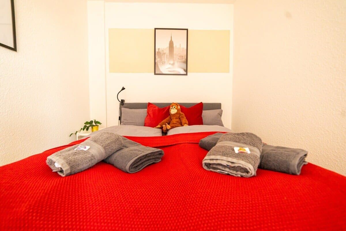 Una cama bien tendida con un edredón rojo, un peluche y dos toallas enrolladas.