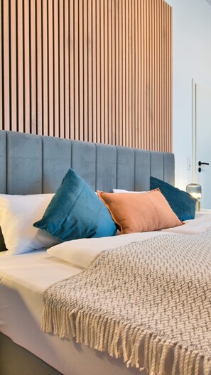 Un lit avec une tête de lit rembourrée de couleur grise, plusieurs coussins dans les tons de bleu et de pêche, et un plaid tricoté de couleur beige.