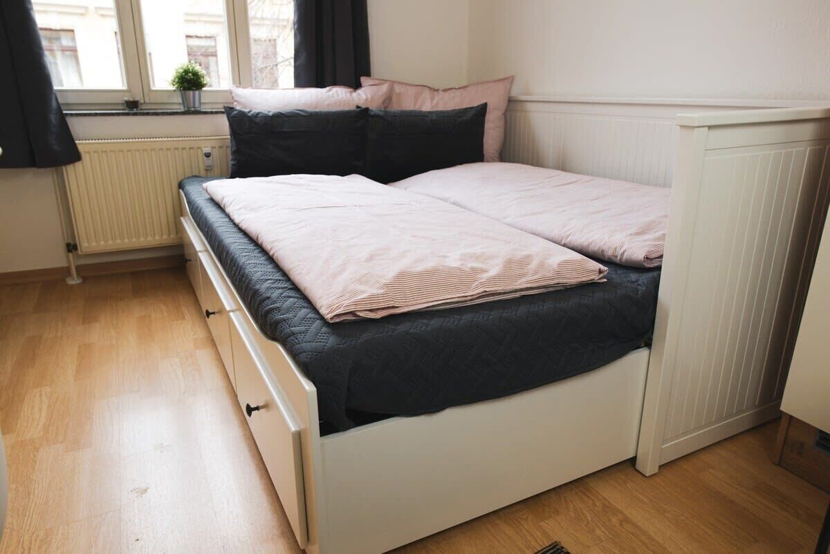 A bedroom with a bed, a radiator, a window with curtains, and a small plant on the windowsill.