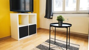 Een kamer met een televisie op een witte standaard, een klein rond tafeltje met een plant, en een raam met gordijnen.