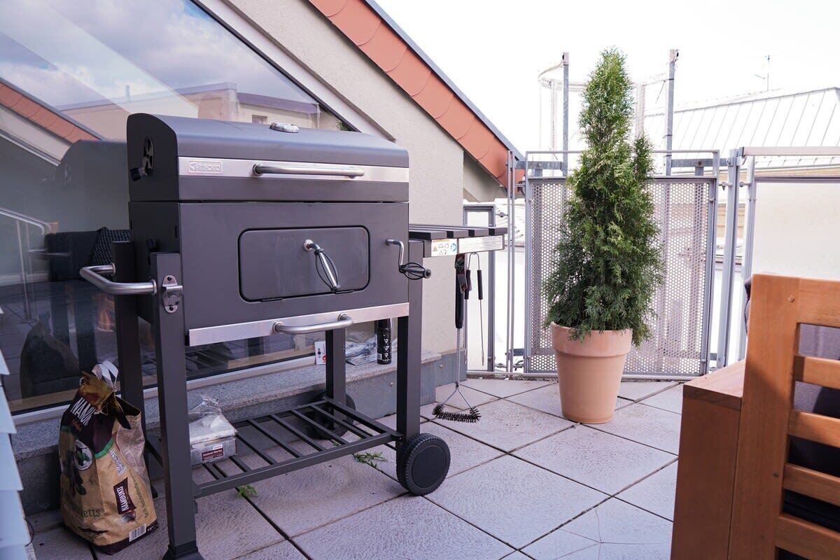 A portable grill on a balcony with a potted plant and a wooden chair.