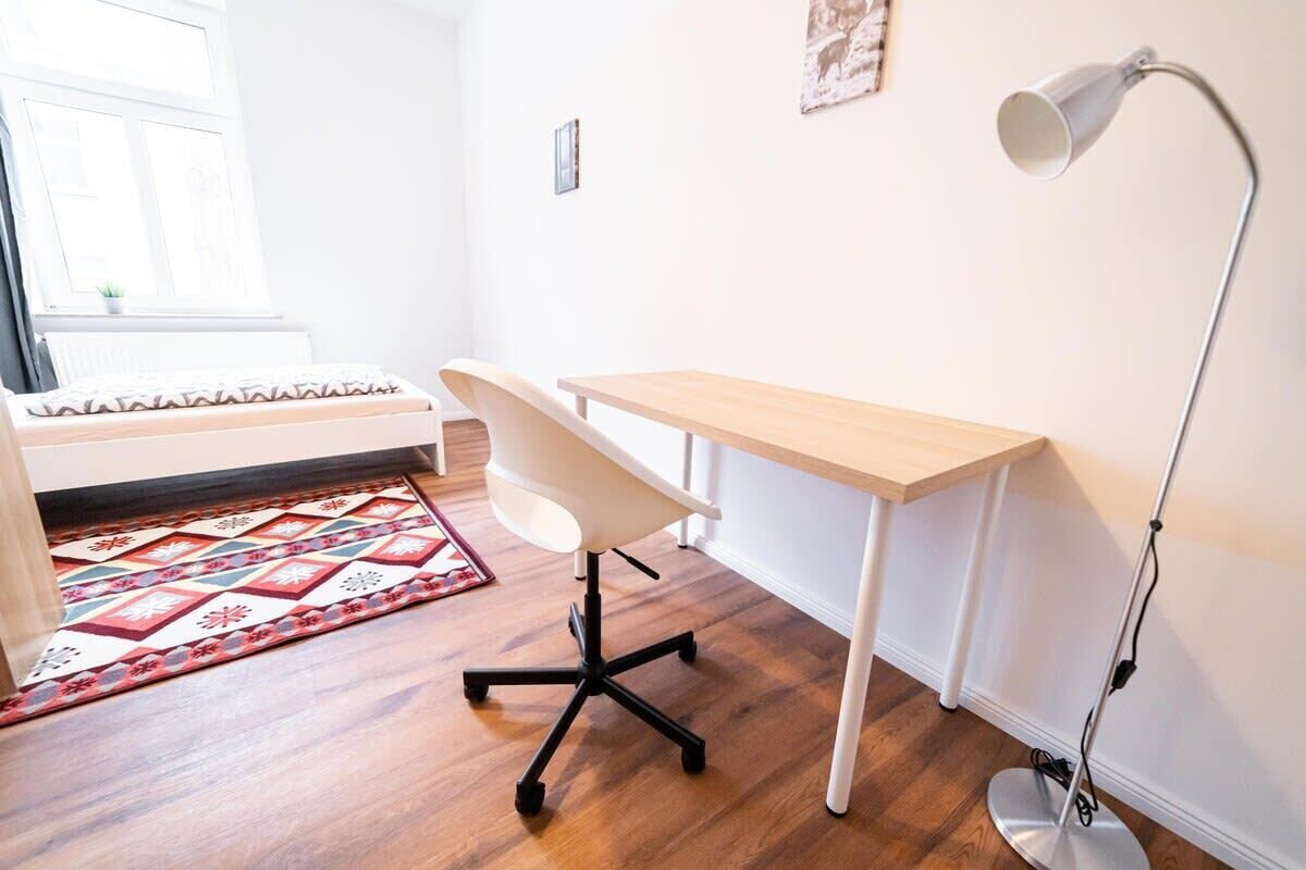 Ein Zimmer mit Holzboden, einem weißen Schreibtisch mit Stuhl, einer Stehlampe und einem gemusterten Teppich.