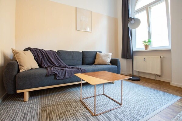 Una sala de estar moderna con un sofá gris, una mesa de centro de madera, alfombra estampada, un radiador y una ventana con una planta.